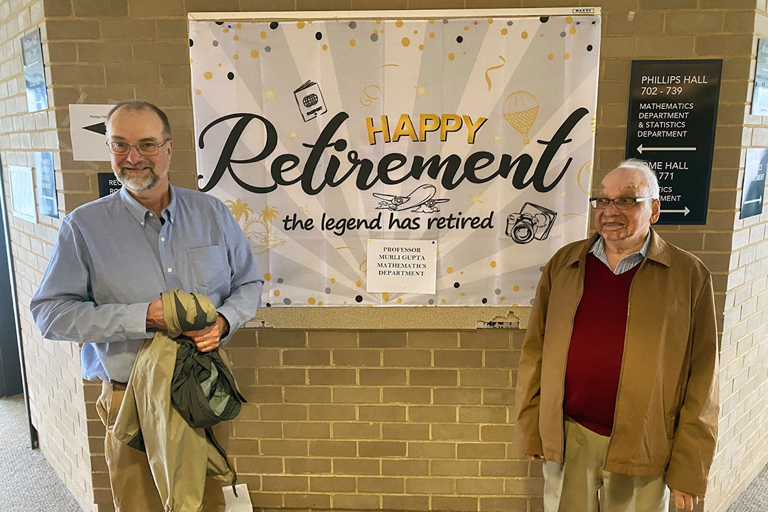 Professors Robbie Robinson and Murli M. Gupta smiling in front of a banner reading Happy Retirement