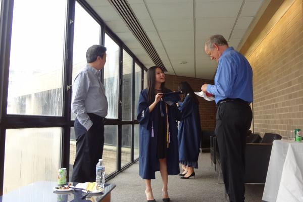 A mathematics graduate wearing a cap and gown talks to two professors at an event
